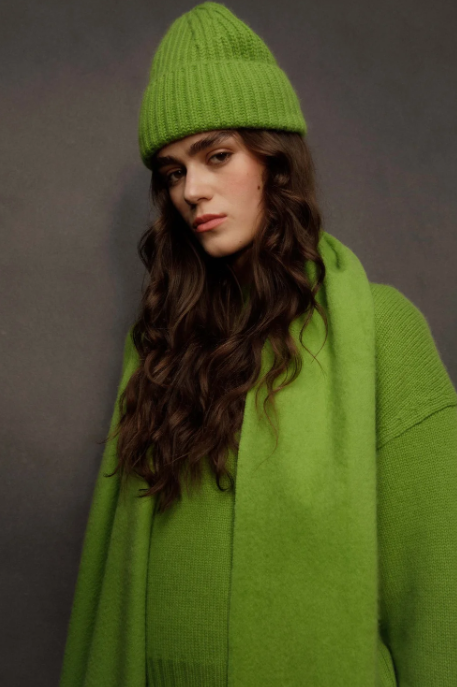 A woman wearing a bright lime green knit beanie, matching knit sweater, and scarf, posed against a dark gray studio background.