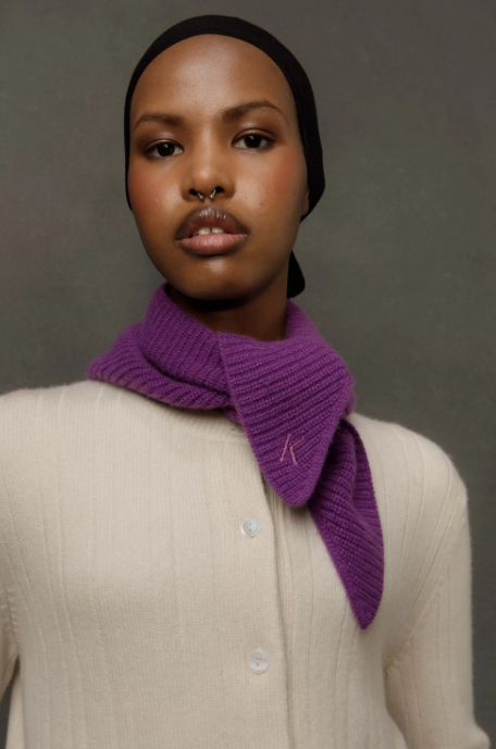 A model wearing a purple knit neck scarf with a small embroidered logo, styled with a cream ribbed cardigan and a black beanie against a neutral gray background.