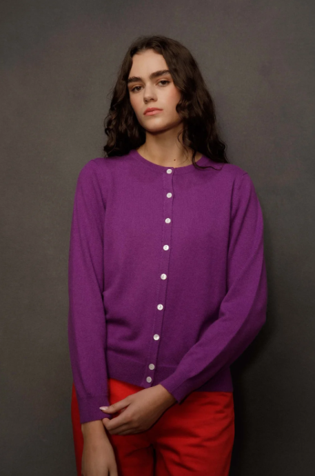 A woman wearing a purple long-sleeve button-front cardigan and bright red pants, standing against a dark gray studio background.