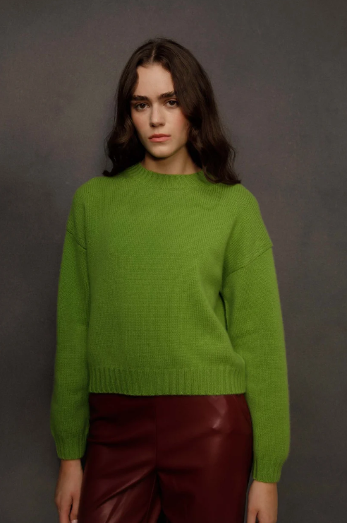 A woman wearing a bright lime green knit crewneck sweater and dark red leather pants, standing against a dark gray studio background.