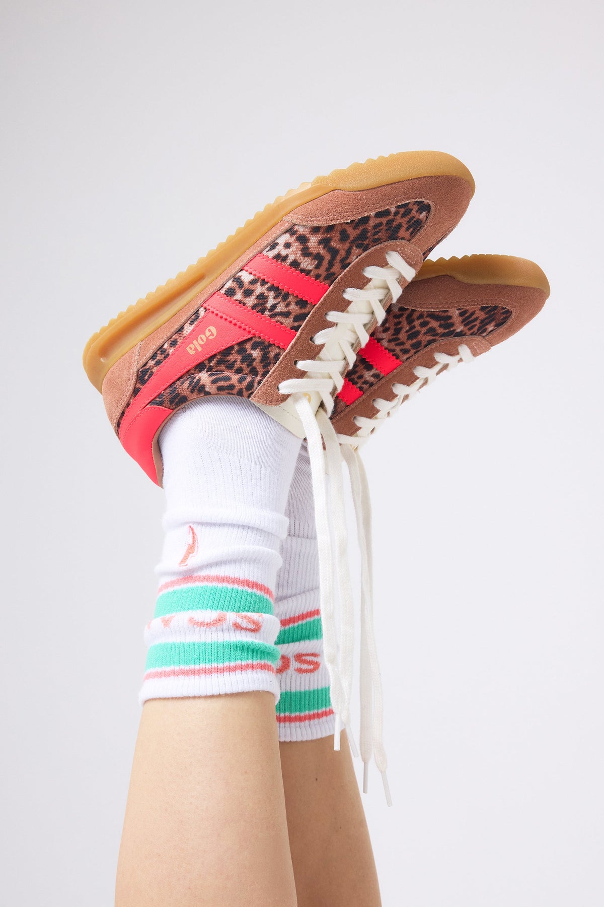 A pair of low-top trainers with leopard print panels, brown suede overlays, and bright pink side stripes, shown worn with white crew socks on a person's legs against a light grey studio background.