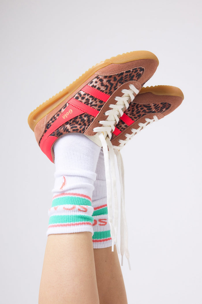 A pair of low-top trainers with leopard print panels, brown suede overlays, and bright pink side stripes, shown worn with white crew socks on a person's legs against a light grey studio background.