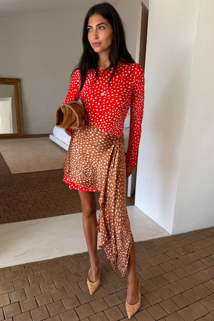 A woman models a red and white polka dot long-sleeve top paired with a brown and white polka dot wrap skirt, holding a brown clutch, in an indoor setting with a tiled floor.