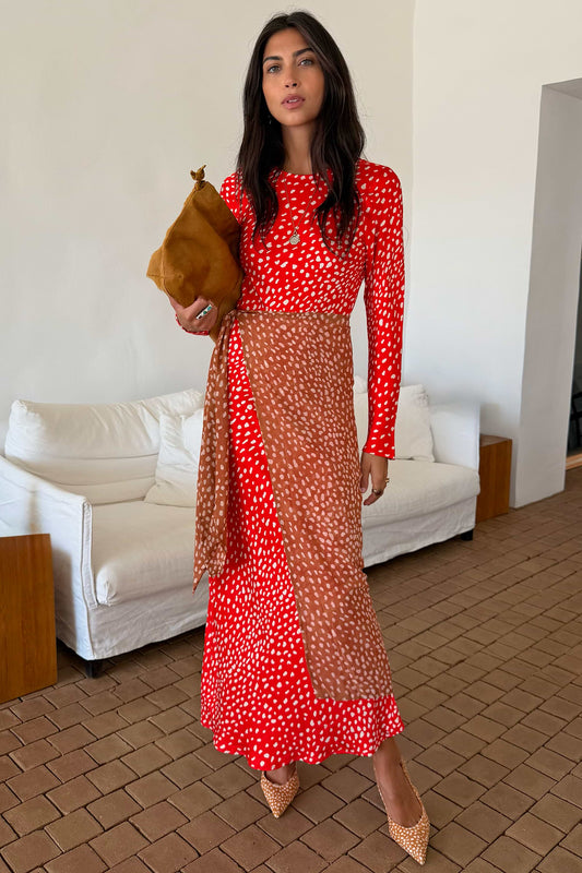 A woman models a red and white patterned maxi dress with a sheer brown mesh overlay, holding a brown clutch bag in an indoor setting with a white sofa in the background.