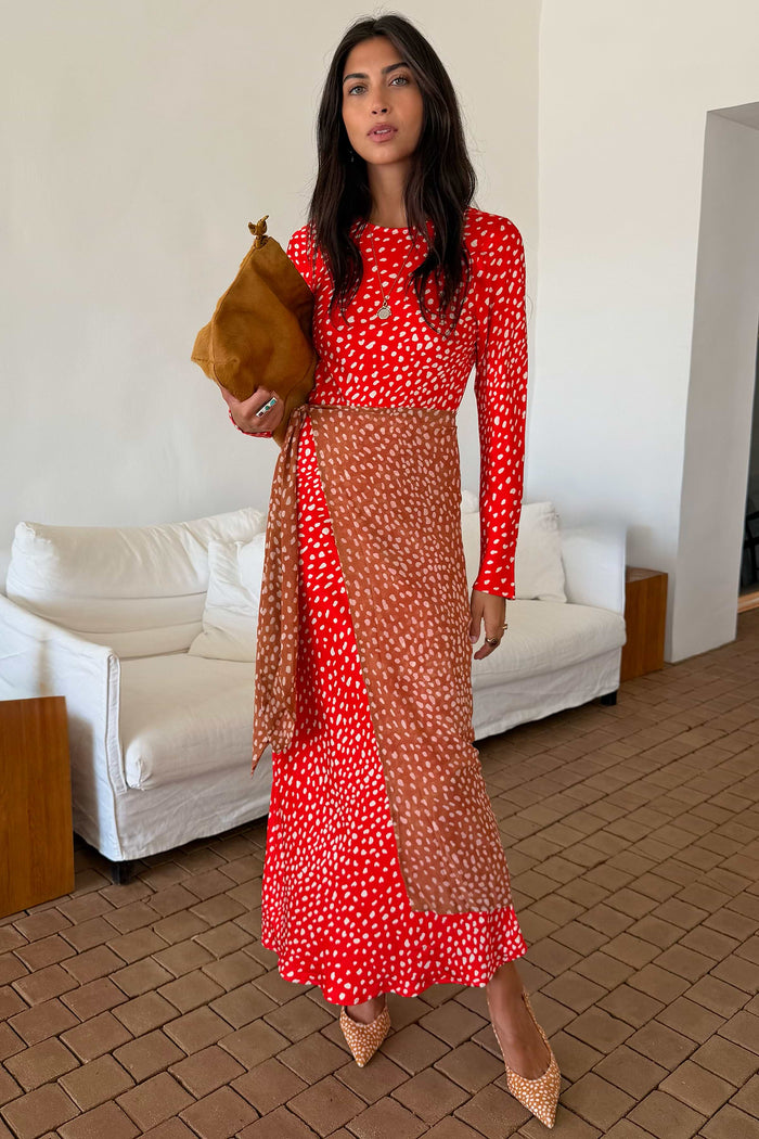 A woman models a red and white patterned maxi dress with a sheer brown mesh overlay, holding a brown clutch bag in an indoor setting with a white sofa in the background.