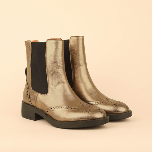 A pair of metallic gold leather Chelsea boots with black elastic side panels and black soles, photographed on a solid light beige background.