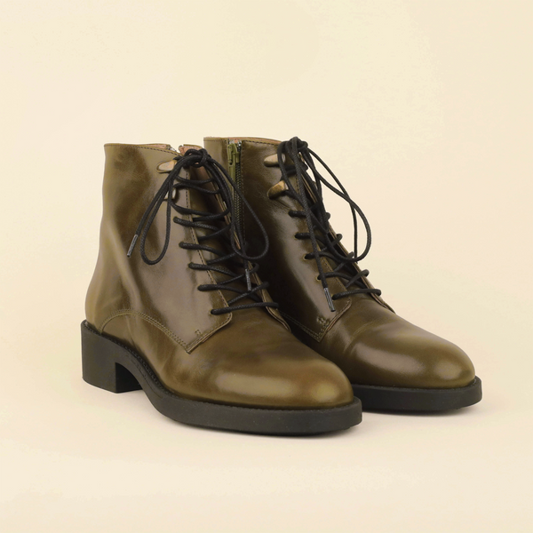 A pair of olive green leather lace-up ankle boots with black laces and black soles, photographed on a clean, solid, warm off-white background.