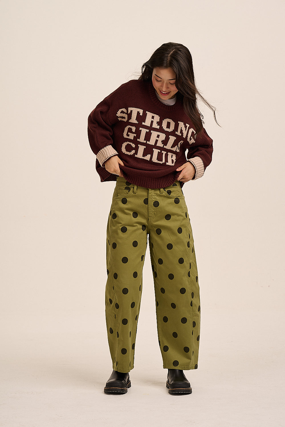 A woman modeling a dark maroon knit sweater with 'STRONG GIRL CLUB' in cream lettering, paired with olive green pants featuring a black polka-dot pattern, against a solid light beige background.