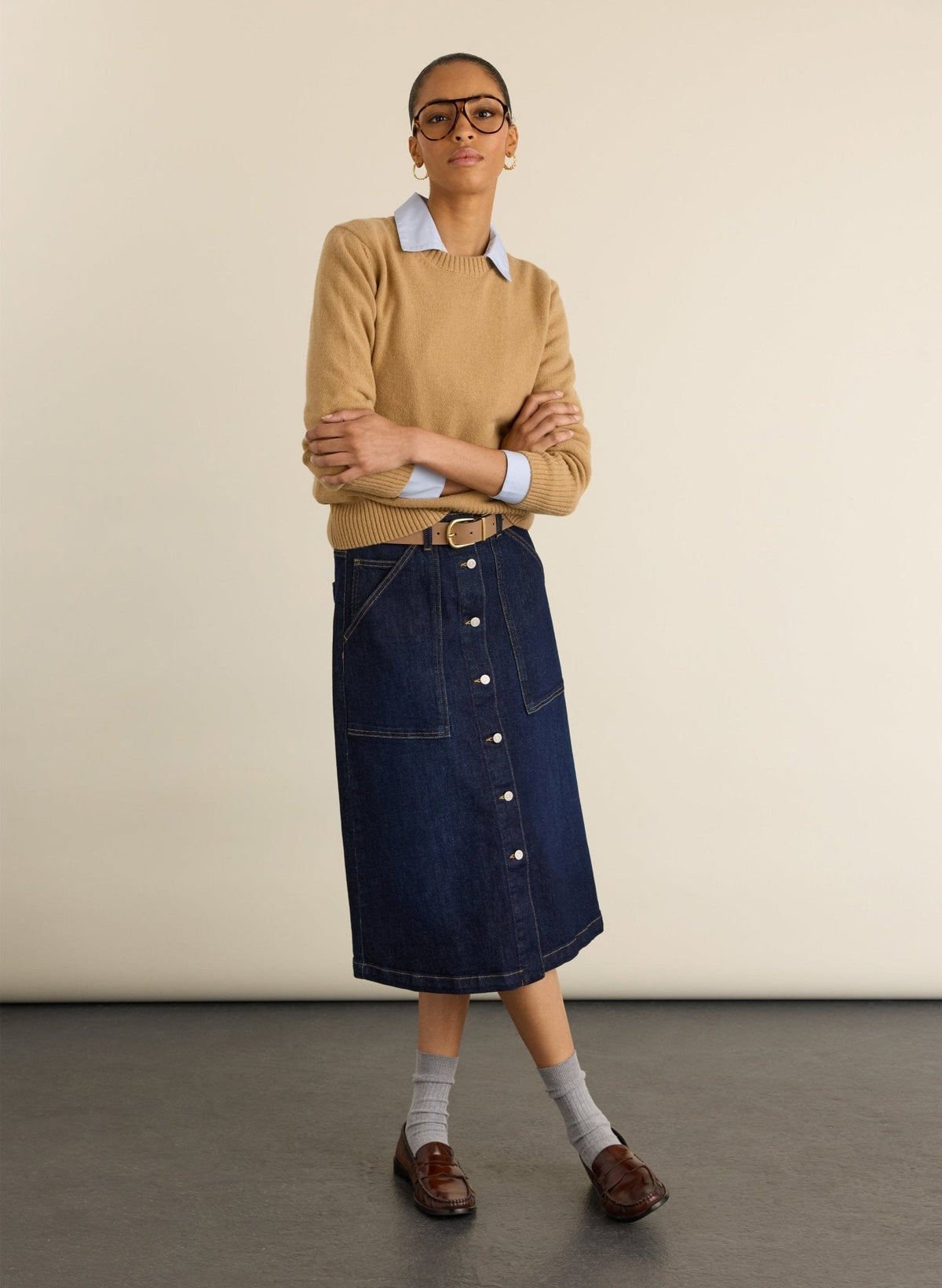 A model stands in a studio wearing a tan sweater over a light blue collared shirt, paired with a dark blue denim button-front skirt, grey socks, brown loafers, and glasses.