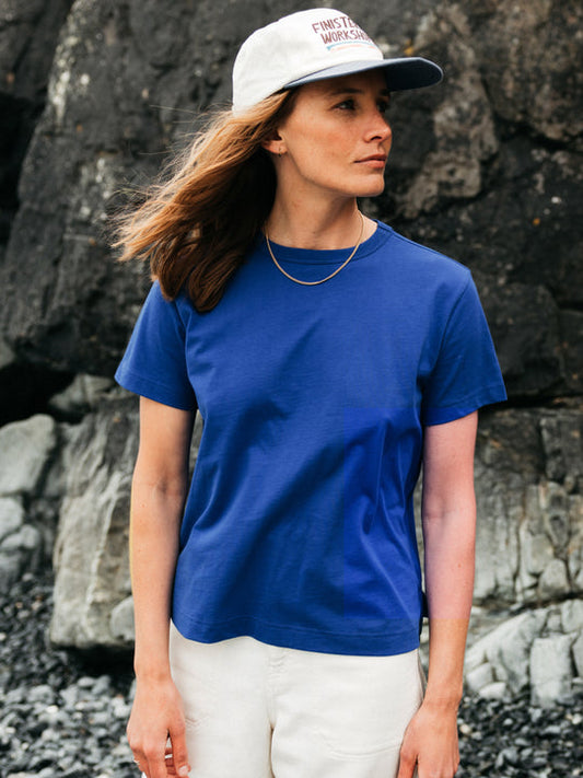 A woman wearing a royal blue short-sleeve crewneck t-shirt, off-white pants, and a white baseball cap with red text, standing outdoors against a dark rocky background.