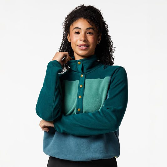 A woman with curly dark hair wearing a color-blocked fleece pullover. The top features a dark teal mock neck and sleeves, a lighter teal chest panel with a button closure, and a muted blue lower section.