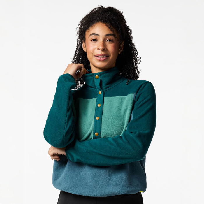 A woman with curly dark hair wearing a color-blocked fleece pullover. The top features a dark teal mock neck and sleeves, a lighter teal chest panel with a button closure, and a muted blue lower section.