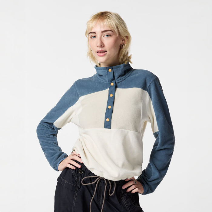 A woman modeling a color-blocked long-sleeve mock neck top with a blue and cream color-blocked design, featuring a button placket with gold buttons, paired with dark drawstring pants against a plain white studio background.