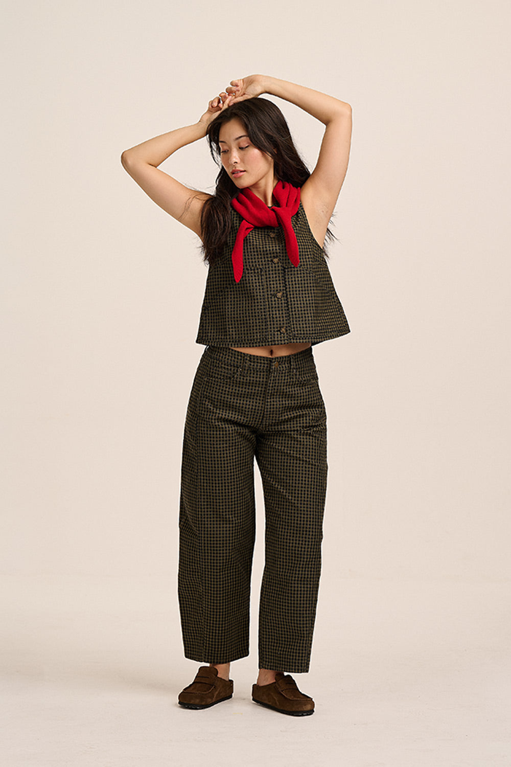 A woman modeling a matching two-piece outfit consisting of a dark olive green and black checkered sleeveless crop top and wide-leg cropped pants, styled with a bright red neck scarf and brown suede shoes, against a solid light beige background.