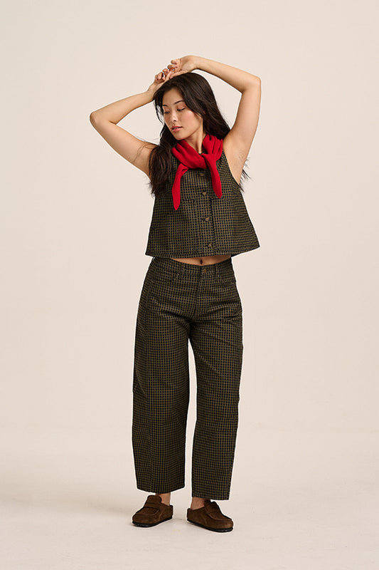 A woman modeling a matching two-piece outfit consisting of a dark olive green and black checkered sleeveless crop top and wide-leg cropped pants, styled with a bright red neck scarf and brown suede shoes, against a solid light beige background.