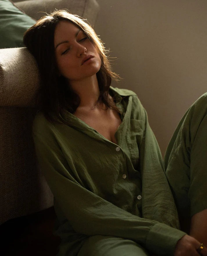 Model sitting against the arm of a sofa, wearing an olive green 100% organic cotton midi shirt pyjama by Nudea, filtered for the Warm Spring, Warm Autumn, and Soft Autumn colour seasons by Shop My Palette.