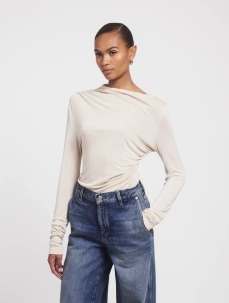A woman modeling a beige long-sleeve top with an asymmetrical draped neckline and ruched side detail, paired with blue jeans, against a plain light gray studio background.
