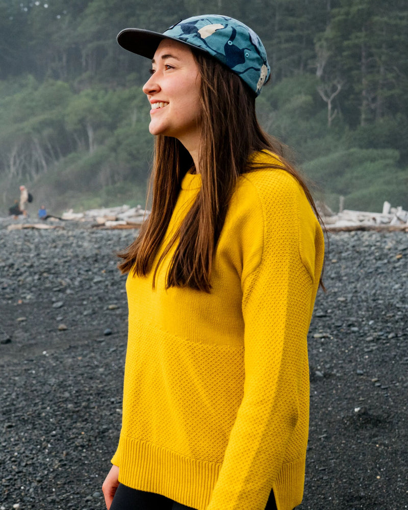 A woman wearing a bright yellow knit sweater and a blue camouflage baseball cap stands on a dark pebble beach with a forested hillside in the background.