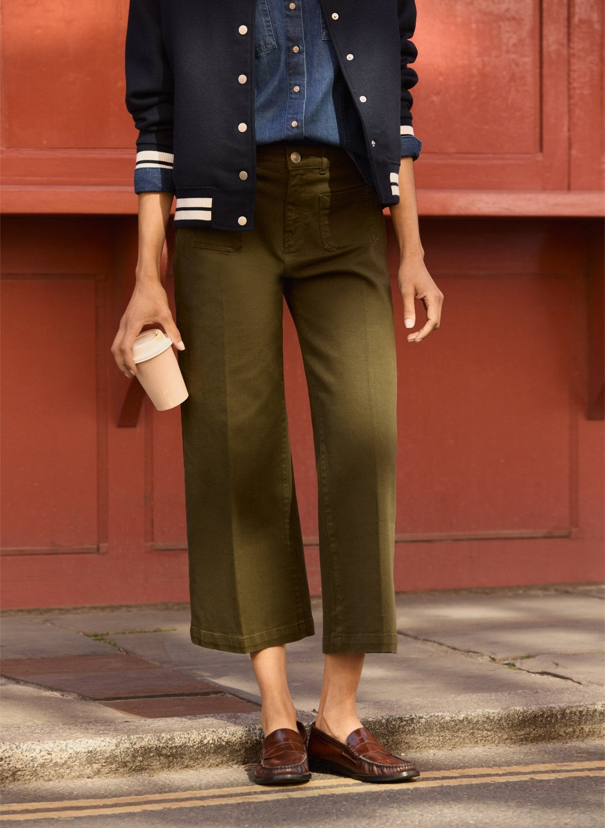 Person wearing a navy jacket with white buttons, blue shirt, dark olive green jeans, and brown shoes against a red wall.