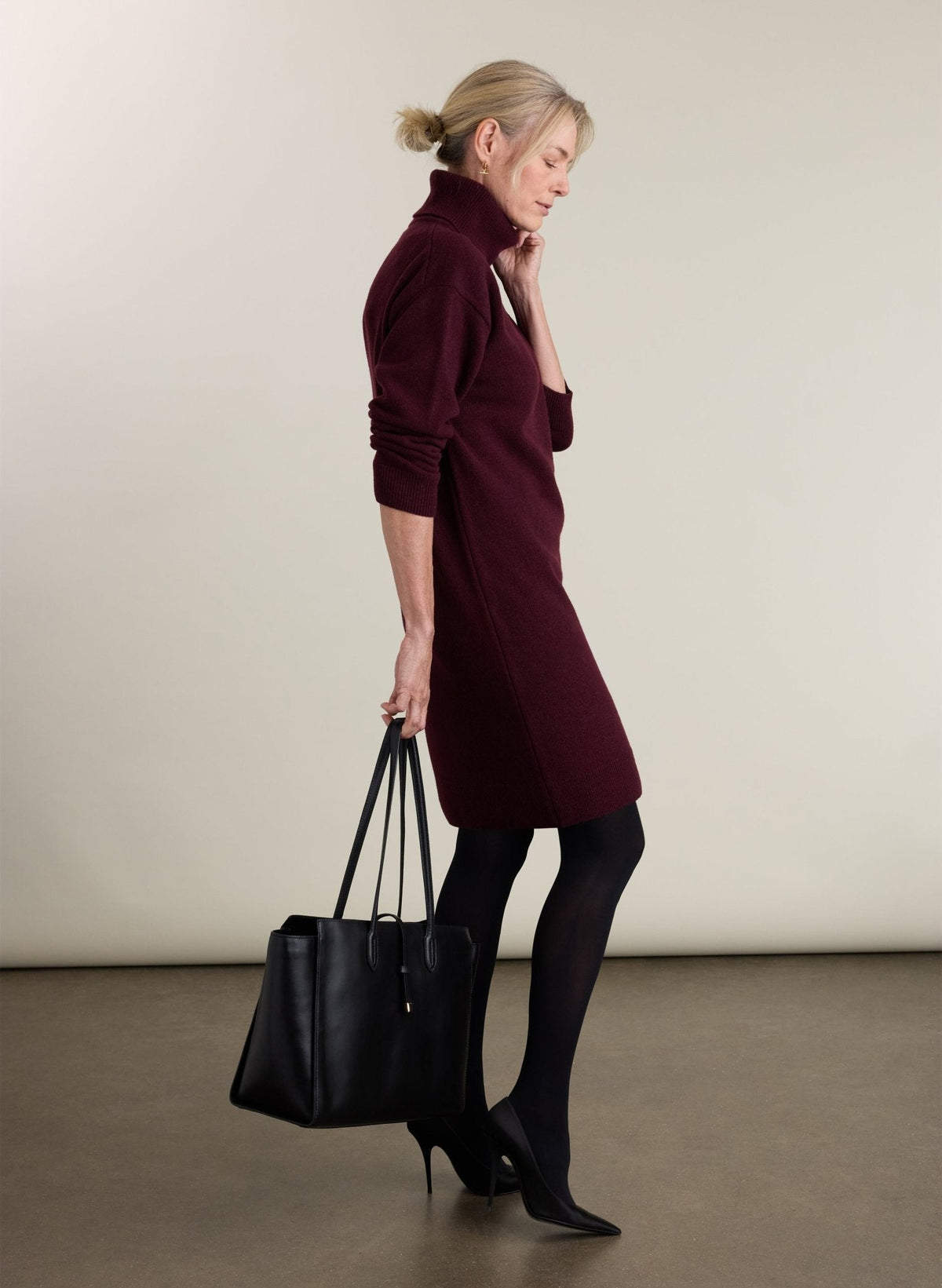 A woman in a side profile wearing a burgundy turtleneck sweater dress, black tights, and black pointed-toe heels, holding a black leather tote bag against a neutral studio background.