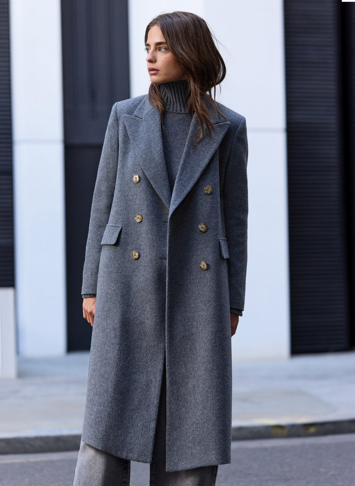 A woman wearing a long double-breasted grey wool coat with buttons over a dark grey turtleneck sweater, standing on a city street.
