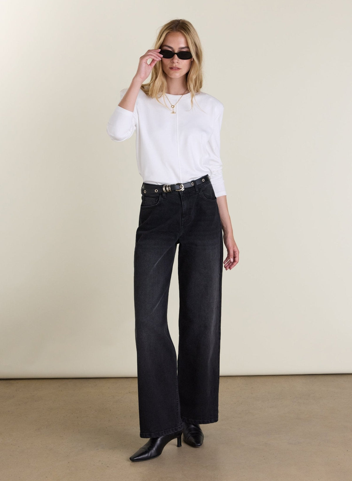 A woman modeling a white long-sleeve top and black wide-leg jeans with a black belt, styled with black pointed-toe heels and sunglasses, standing against a plain beige studio backdrop.