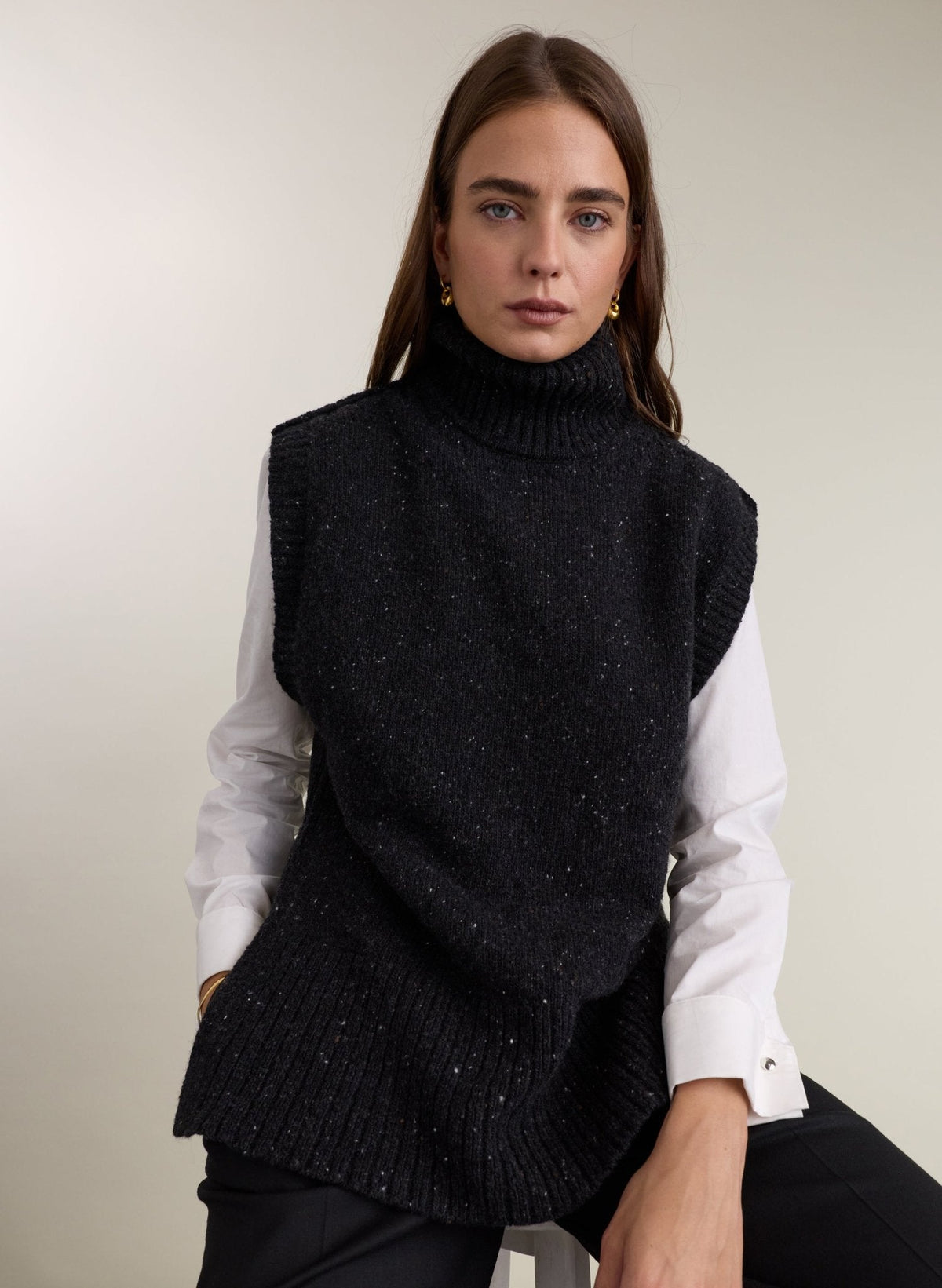 A woman wearing a charcoal marl knit turtleneck sweater vest over a white long-sleeve button-up shirt, photographed against a plain light beige studio background.
