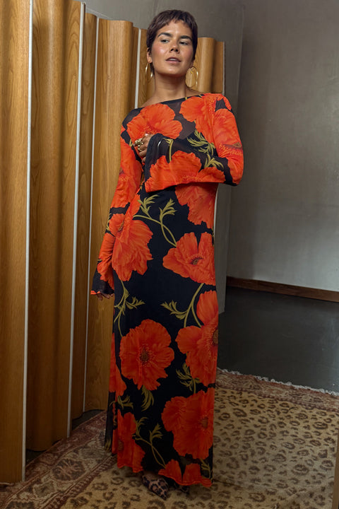 A woman wearing a long-sleeved maxi dress with a bold black and orange floral print, standing in front of wooden folding screens and a patterned rug.