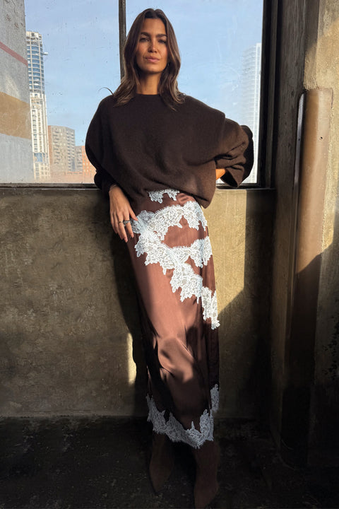 Woman wearing a brown sweater and lace skirt standing against a cityscape background