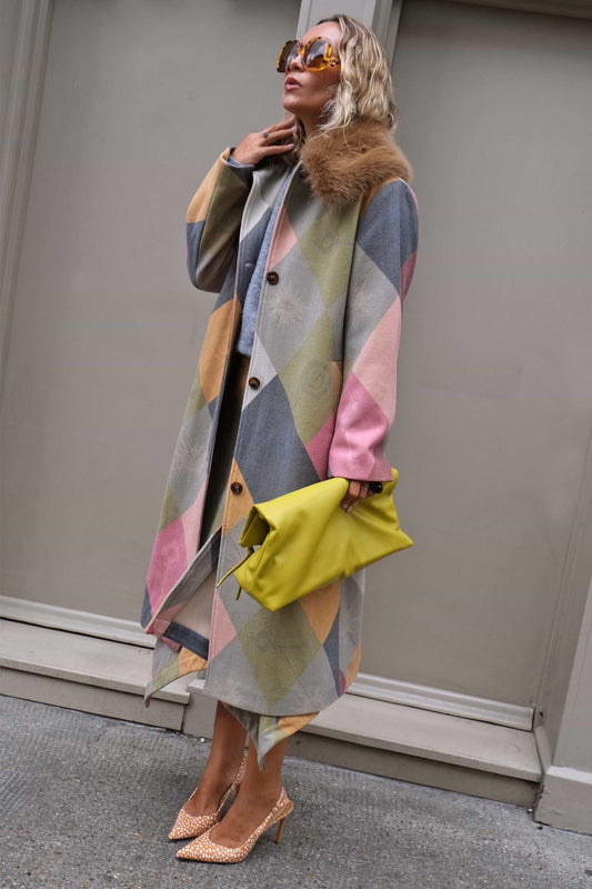 A woman models a long geometric print coat with a brown fur collar, holding a bright yellow clutch and wearing leopard-print heels against a gray building wall.