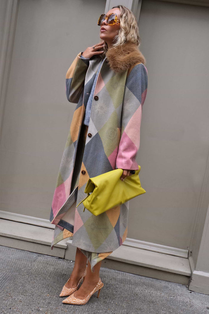 A woman models a long geometric print coat with a brown fur collar, holding a bright yellow clutch and wearing leopard-print heels against a gray building wall.
