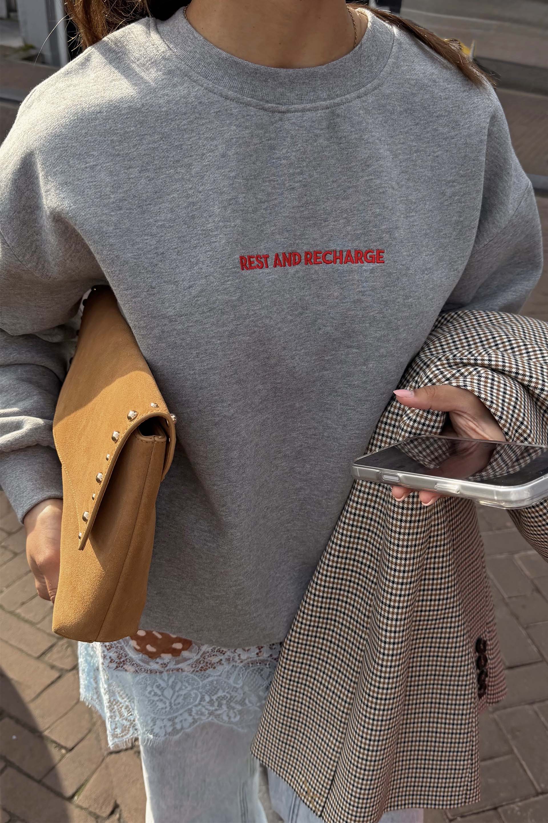 Grey crewneck sweatshirt with small red embroidered text on the chest reading “Rest and Recharge.”