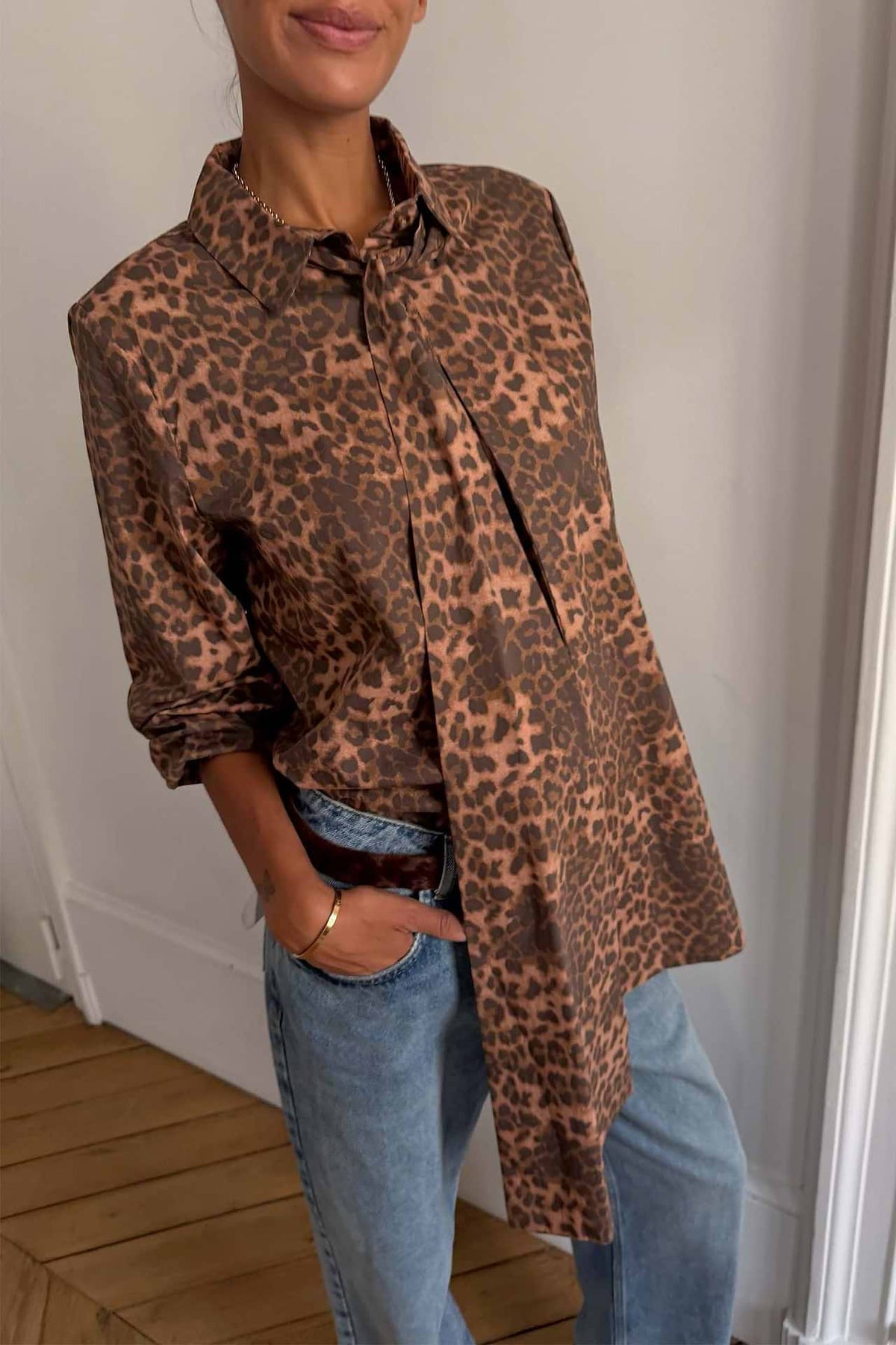 A woman wearing a brown and beige leopard-print button-up shirt with rolled sleeves, tucked into light-wash blue jeans, standing indoors against a white wall.