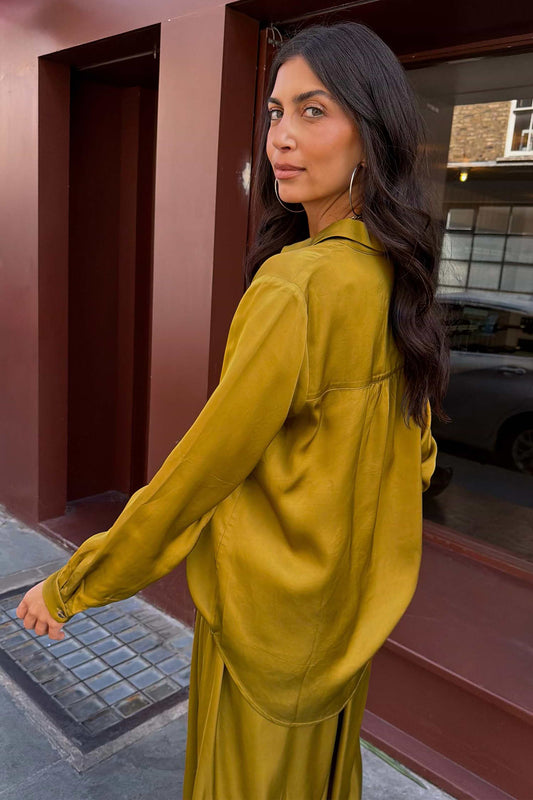A woman wearing a flowing olive yellow satin long-sleeve top and matching skirt, photographed outdoors in front of a dark red-brown building.