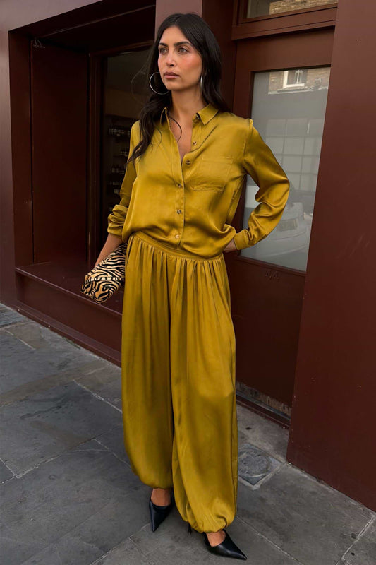 A woman stands on a stone pavement in front of a dark brown building, wearing a mustard yellow satin long-sleeve button-up shirt and matching wide-leg trousers. She is holding a tiger-print clutch and wearing black pointed-toe heels.