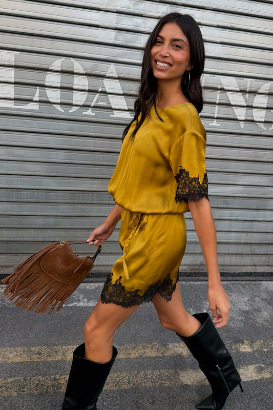 A woman wearing a mustard yellow t-shirt dress with black lace trim walks in front of a grey metal shutter. She is holding a brown fringe handbag and wearing black knee-high boots.