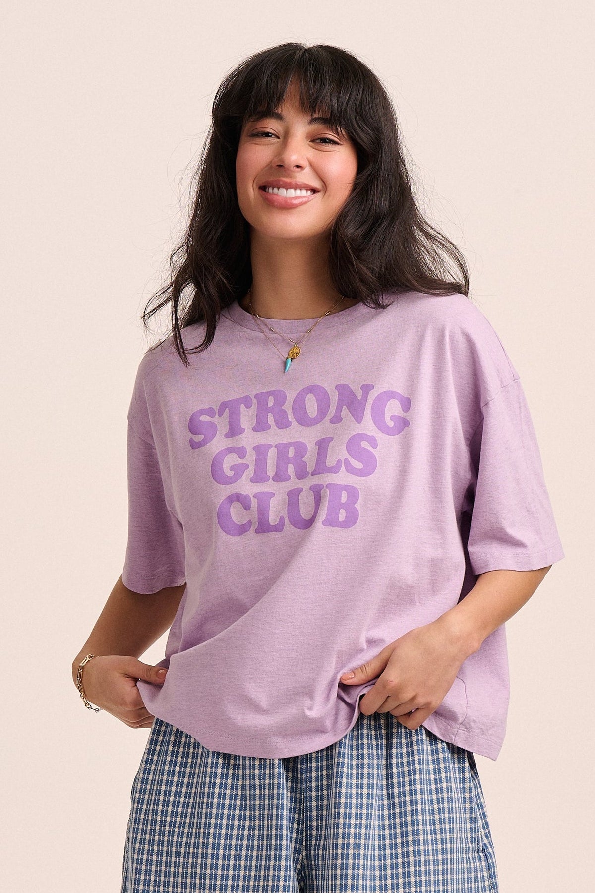 Woman wearing a purple 'STRONG GIRLS CLUB' t-shirt against a beige background