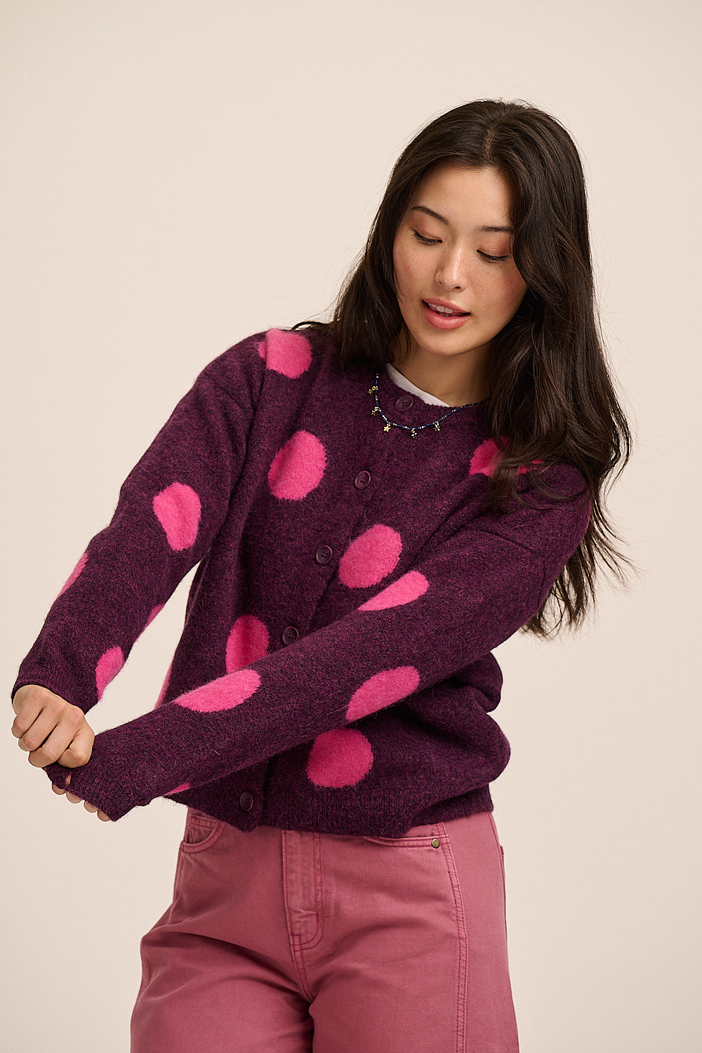 A woman wearing a dark purple fuzzy cardigan with large hot pink polka dots, paired with dusty rose pants, against a plain beige background.