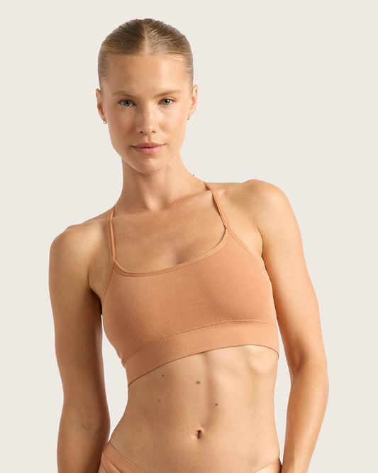 A woman modeling a seamless, scoop-neck sports bra in a warm peach-beige color against a solid light gray background.
