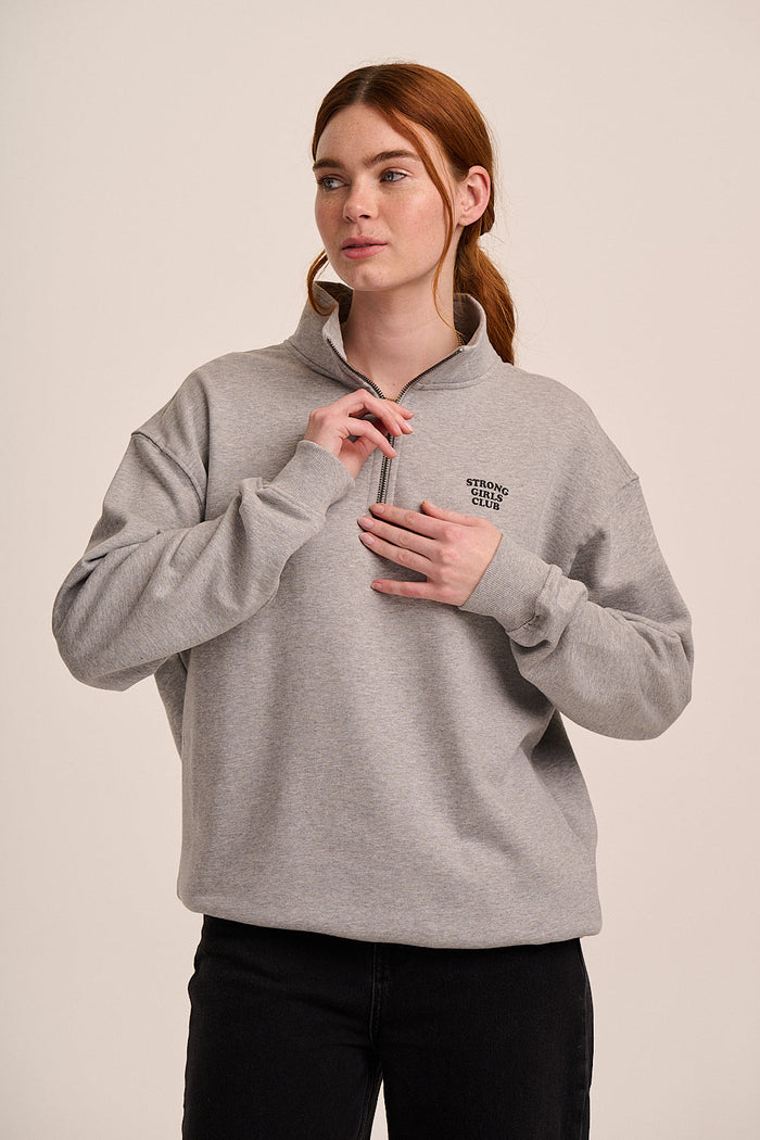 Mid grey half-zip sweatshirt with high collar, small ‘Strong Girls Club’ logo, long sleeves, and relaxed fit.