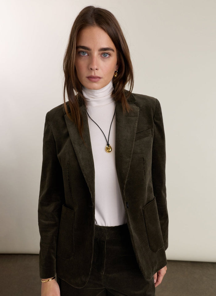 A woman wearing a dark olive green corduroy blazer over a white turtleneck sweater, paired with dark brown corduroy trousers and a gold pendant necklace, standing against a plain off-white studio background.