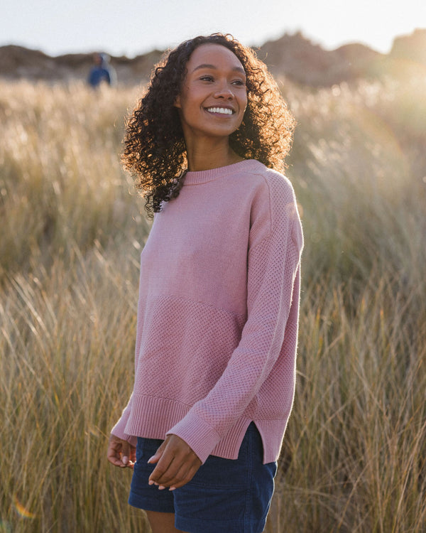 A smiling woman stands outdoors in a field of dry grass, wearing a pink textured knit sweater and dark blue shorts.